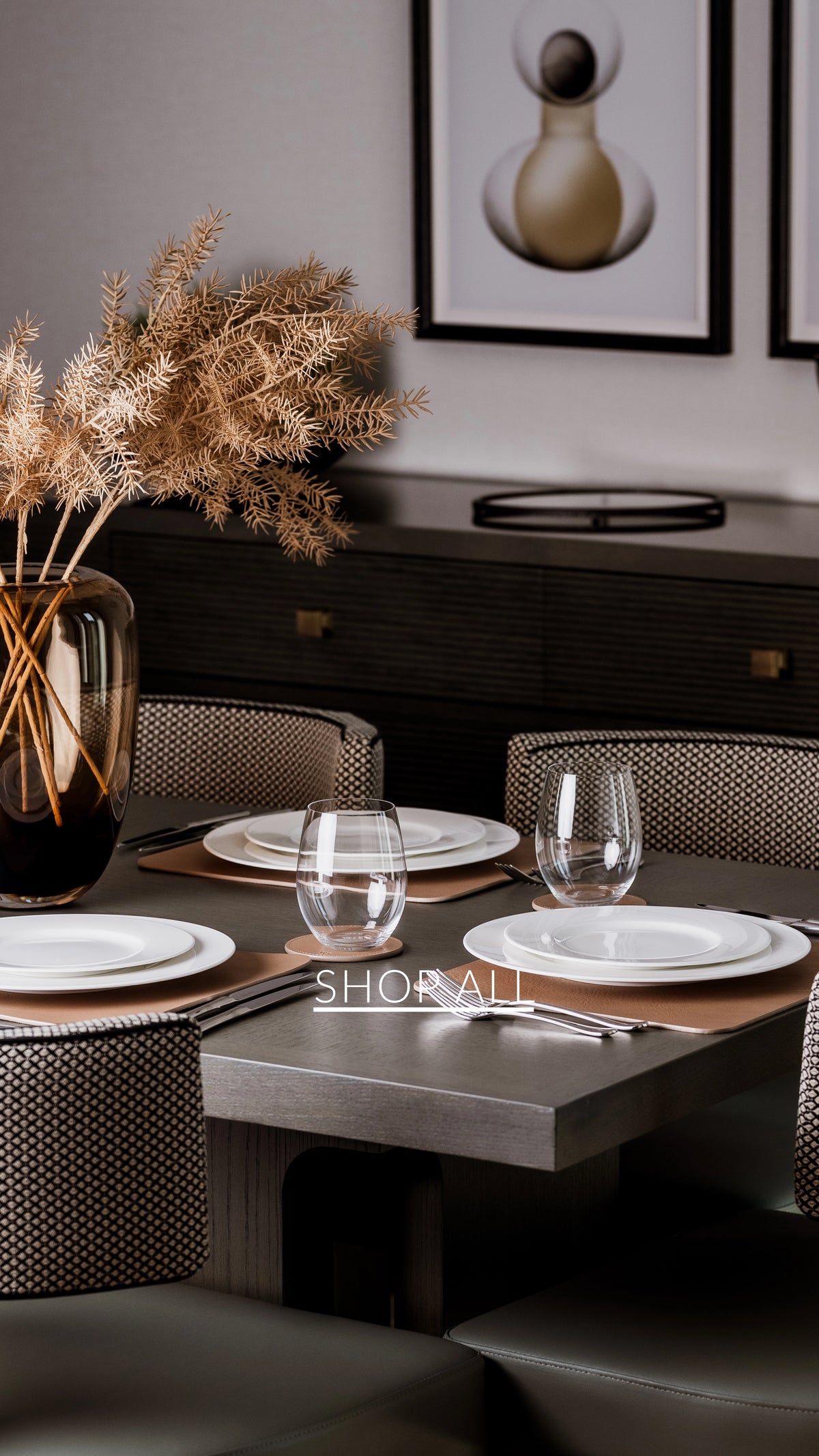 Dining table setting with plates, glasses, and a vase of dried plants in a modern interior.