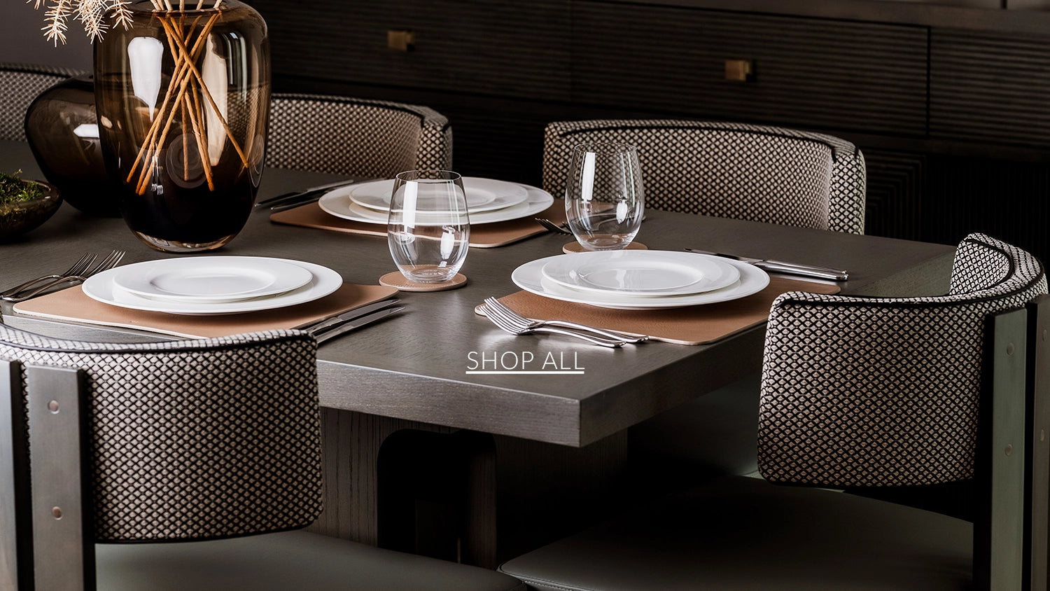 Dining table set with plates, glasses, and cutlery on a dark surface with chairs around it.